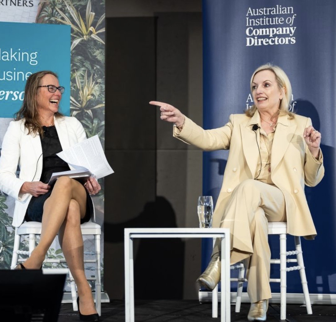 Jenny sat on a chair during a panel discussion, smiling, with Christine Holgate, a fabulous female trailblazing CEO who was inappropriately sacked by the PM and went on to run the Government competitor. Jenny interviewed her for the Australian Institute of Company Directors and it was an incredibly powerful and purposeful interview for women. Both are wearing a suit, and Jen laughs whilst Christine points into the audience.