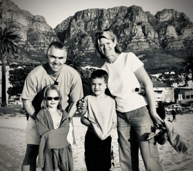 A black-and-white picture of Charles with Kris and their two children on a beach in Cape Town, with rocky hills in the background