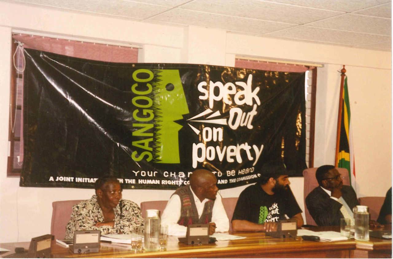 The picture shows Kumi speaking on a panel. The banner behind them reads 'Speak out on poverty'.