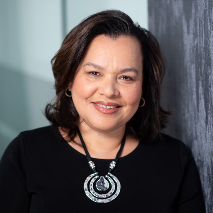 Headshot of Lisette, wearing a black dress and a necklace with a large silver pendant. 