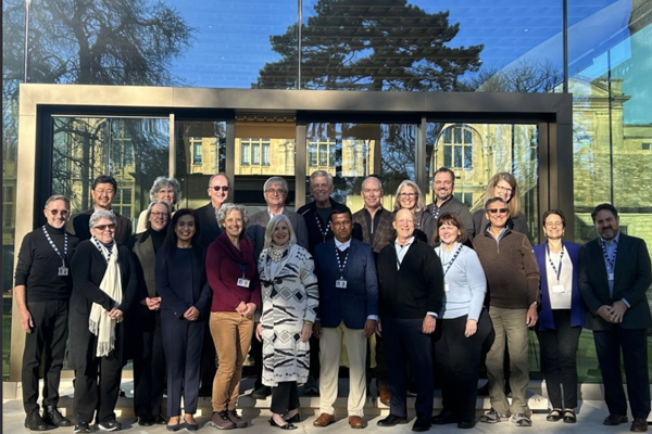 A group of twenty Oxford Next Horizons participants wearing lanyards, posing in the sunshine. They are standing outside the doors of the Rhodes garden pavillion, which is a large, reflective, glass building.