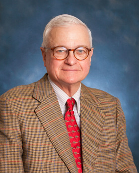 Portrait photo of Peter wearing a tweed suit, red tie, glasses, and a blue background.