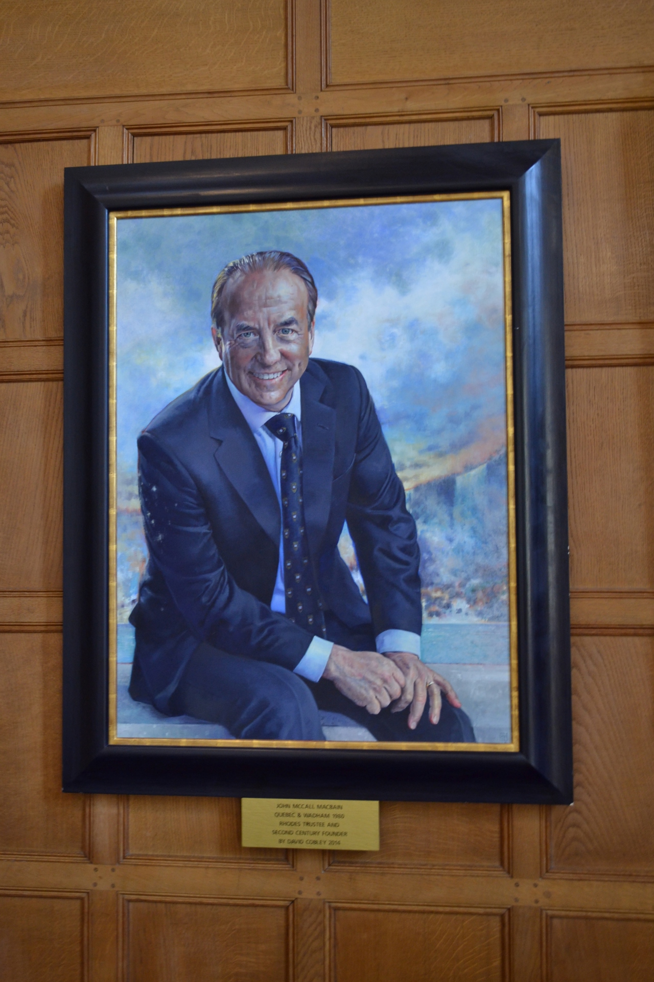 Image of a painting of John, he is sat in a suit, and smiling, with his hands on his legs.