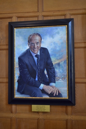 Image of a painting of John, he is sat in a suit, and smiling, with his hands on his legs.
