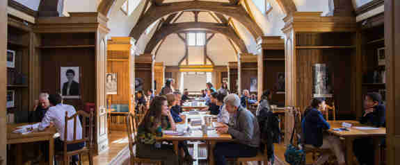 A wide shot of several people sitting at tables in a library. Most are sat in pairs and are in the middle of a discussion.