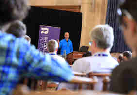 Image of a man in a blue jumper standing at the front of a lecture hall speaking to a room of seated attendees. A sign saying Rhodes Ventures is in the background. 