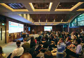 An image of a conference room filled with people listening to three speakers on stage. 