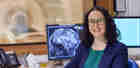 Image of a smiling woman in a blue jacket sitting in front of a computer screen that shows an x-ray scan of a brain.